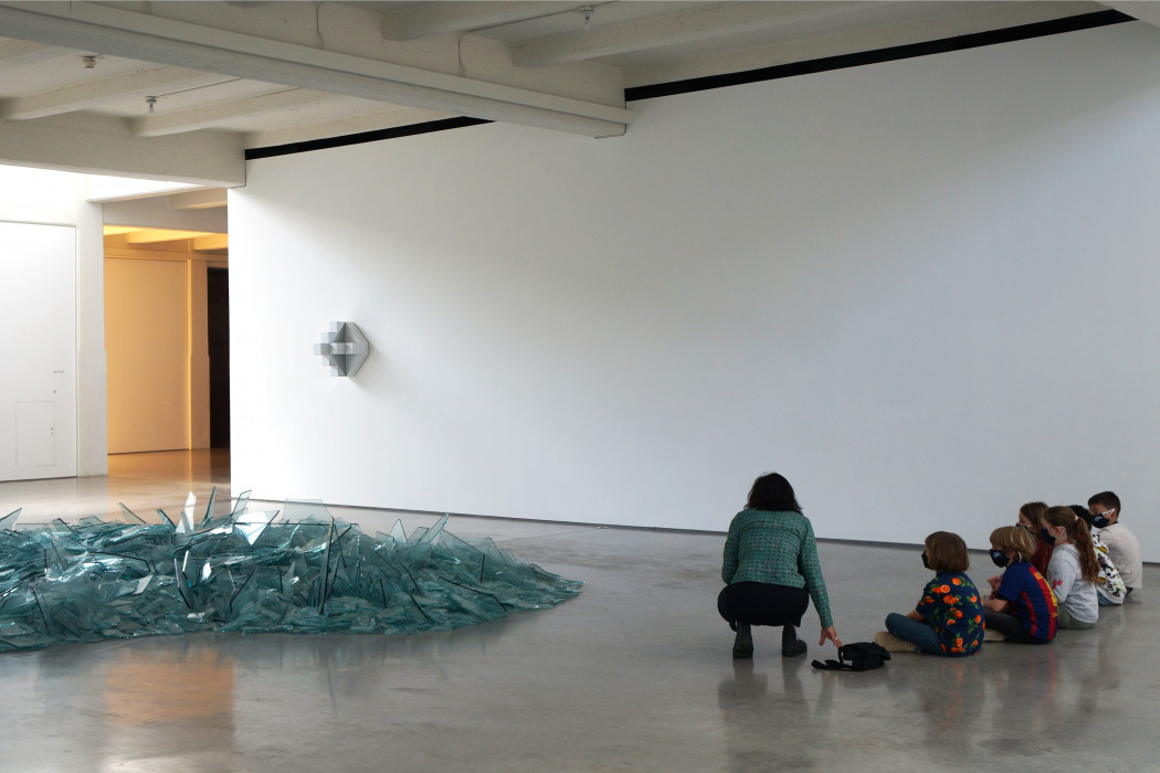 A group of children sit in front of an artwork made from broken glass. Nearby, a teacher is crouched and in the middle of talking to the children.